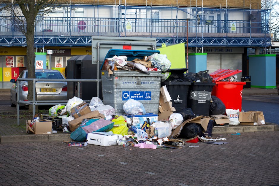 A large collection of mixed waste and rubbish is piled on a paved street within a designated waste collection area, including cardboard boxes, plastic bags filled with refuse, crumpled paper, and vari