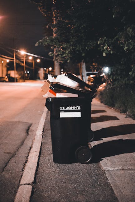 A large collection of mixed waste and rubbish is piled on a paved street within a designated waste collection area, including cardboard boxes, plastic bags filled with refuse, crumpled paper, and vari